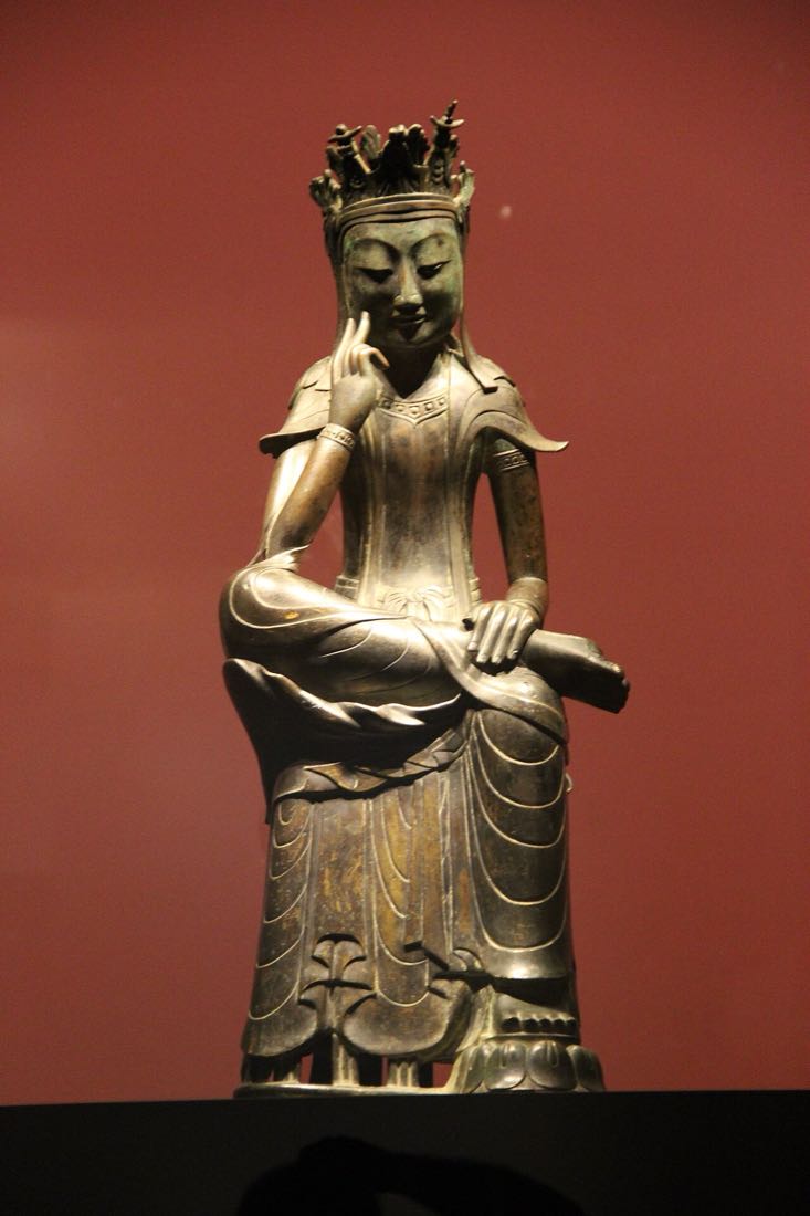 붉은 배경 앞에 한쪽 다리를 올리고 손가락을 뺨에 댄 채 앉은 금동 불상